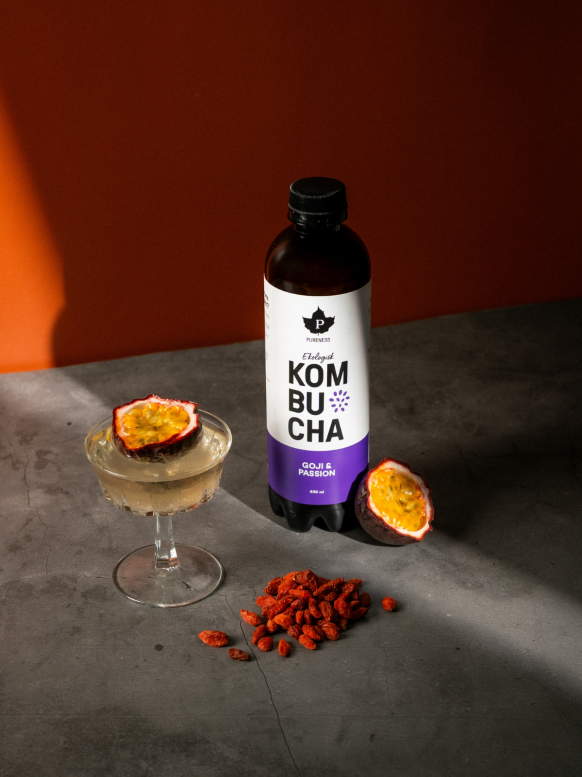 pureness kombucha goji passion flaska bredvid ett glas med kombucha och en passionsfrukt uppskuren i glaset och gojibär framför