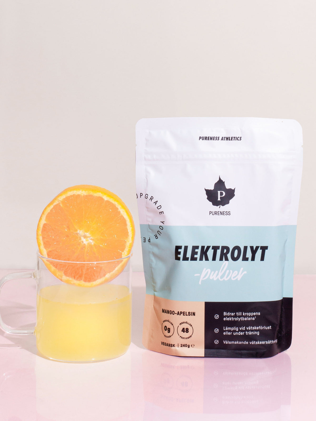 en påse elekotrlytpulver med smal mango-apelsin bredvid en glasmugg med elektrolyt-dricka och en apelsin i 