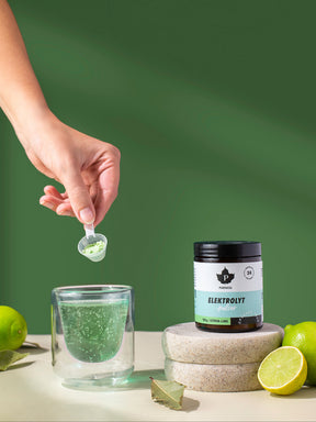 en hand som häller elektrolyter i ett glas bredvid en burk elektrolytpulver citron lime som står på en sten med limes runt om