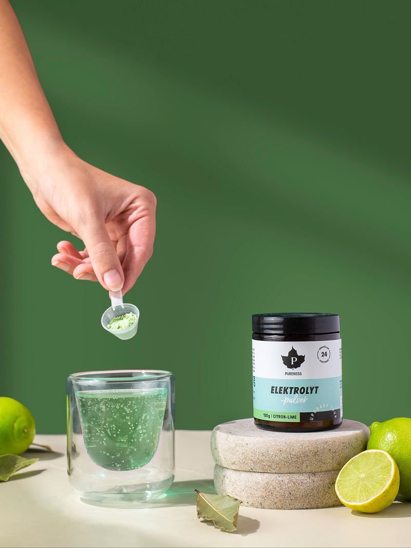 en hand som häller elektrolyter i ett glas bredvid en burk elektrolytpulver citron lime som står på en sten med limes runt om