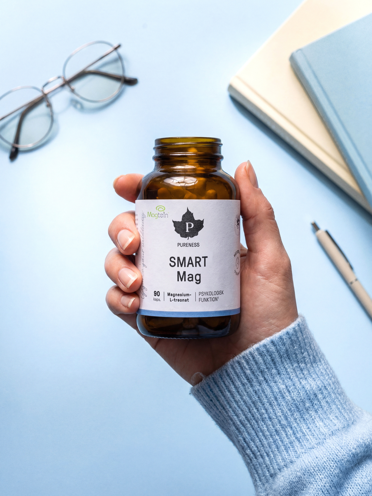 En burk Smart Mag som hålls av en hand med en blå bakgrund med böcker och ett par glasögon