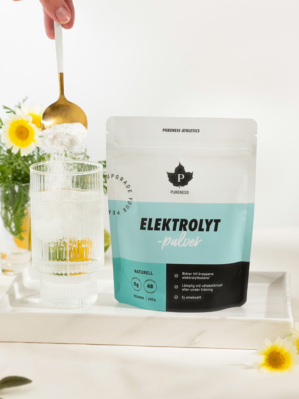 en påse pureness elektrolytpulver bredvid ett glas där elektrolytpulvret hälls i med en vas blommor i bakgrunden