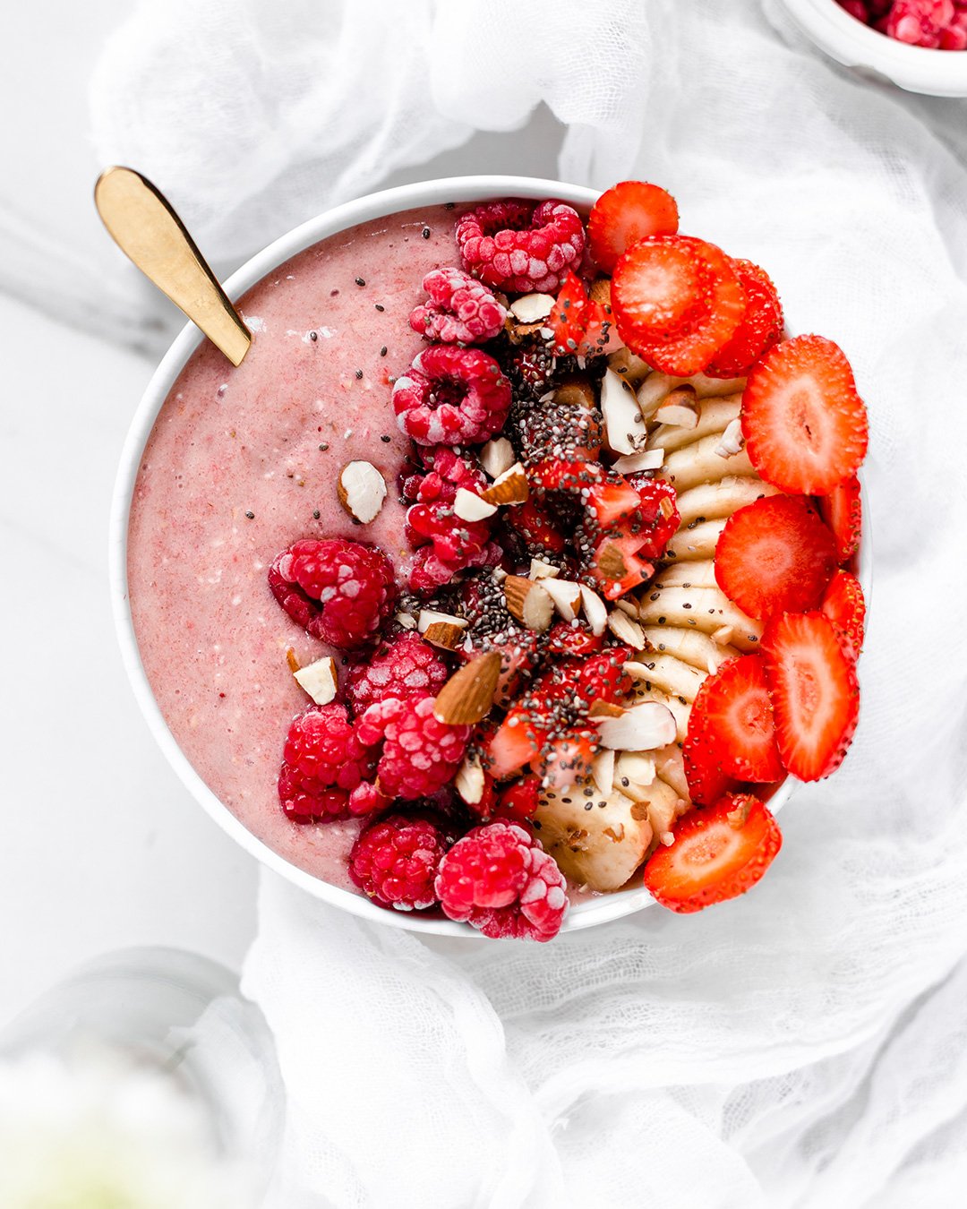 En smoothie bowl med hallon banan och jordgubb på ett bord med vit dekor
